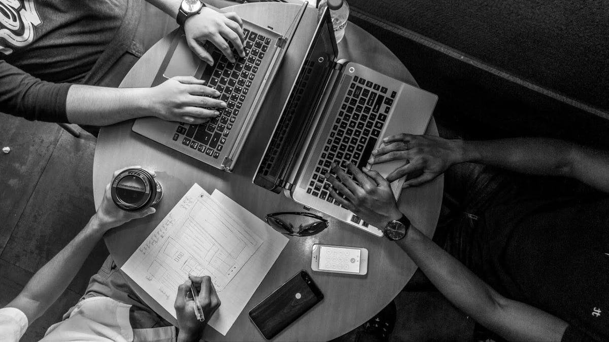 Overhead black and white view of two people collaborating at a desk with laptops, notebooks, coffee, and mobile phones